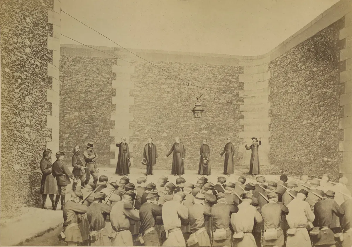 Cour de prison aux hauts murs, six otages debout alignés contre le mur dont plusieurs religieux, face à un peloton de gardes nationaux vus de dos