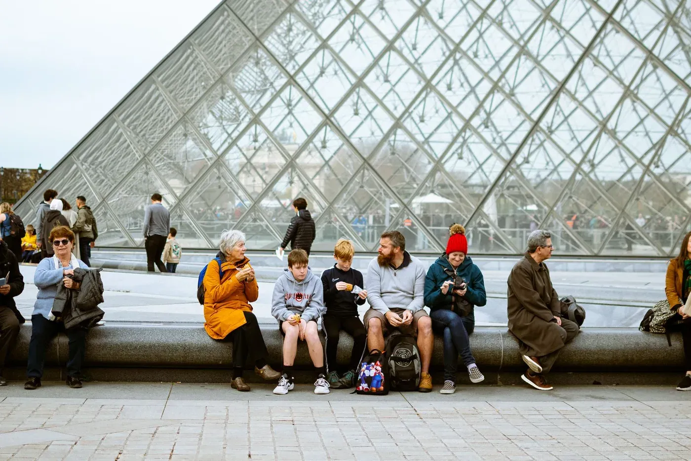 Une famille assise devant la pyramide du Louvre, viennoiseries à la main, avec deux adolescents