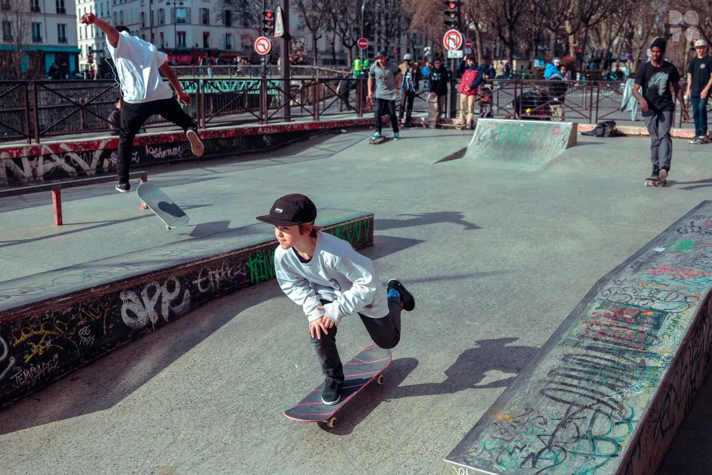 Jeunes skateurs dans un skatepark parisien couvert de graffitis, immeubles haussmanniens en fond